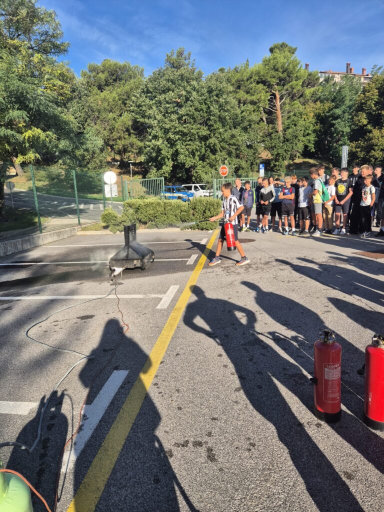 Učenci gasijo s pomočjo simulatorja gasilnega aparata.